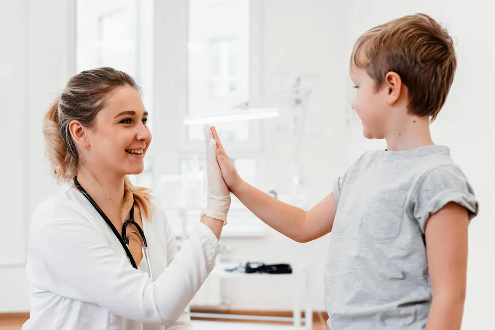 A smiling female paediatrician, seated, high-fives a standing boy in a clinical setting, celebrating a positive and encouraging moment.