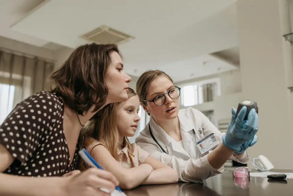A mother, child, and female clinician sit at a table, attentively examining a glucometer. The clinician holds the device while the mother leans in, taking notes with a pen on paper.