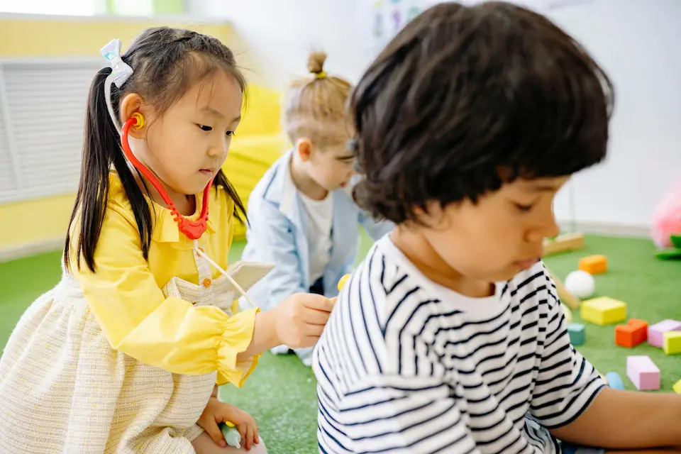 In a daycare or similar setting, three young children engage in different activities. In the foreground, a girl in a bright yellow dress playfully performs a 'check-up' on her classmate using a toy stethoscope, while her patient reads or interacts with something out of frame. In the background, another child plays with blocks, partially obscured.